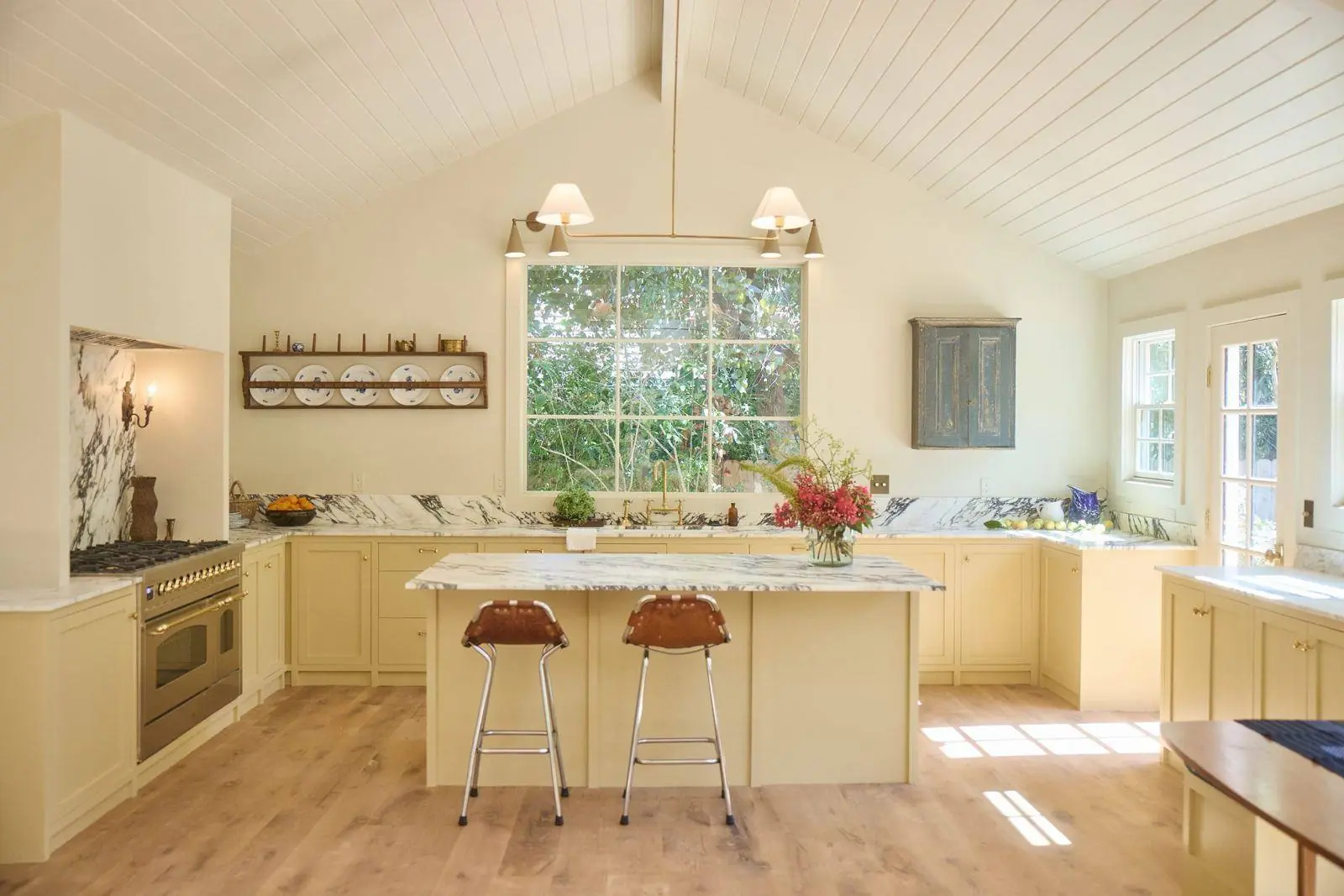 A painted cottage kitchen with a marble-topped island, leather stools and a window over the sink
